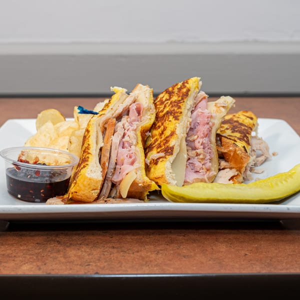 Ida's Cafe breakfast sandwich with ham, cheese, and sauce, served with chips and a pickle.
