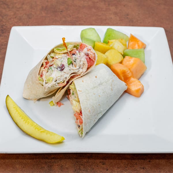 Chicken salad wrap with lettuce and tomato, served with a pickle spear and fruit salad.